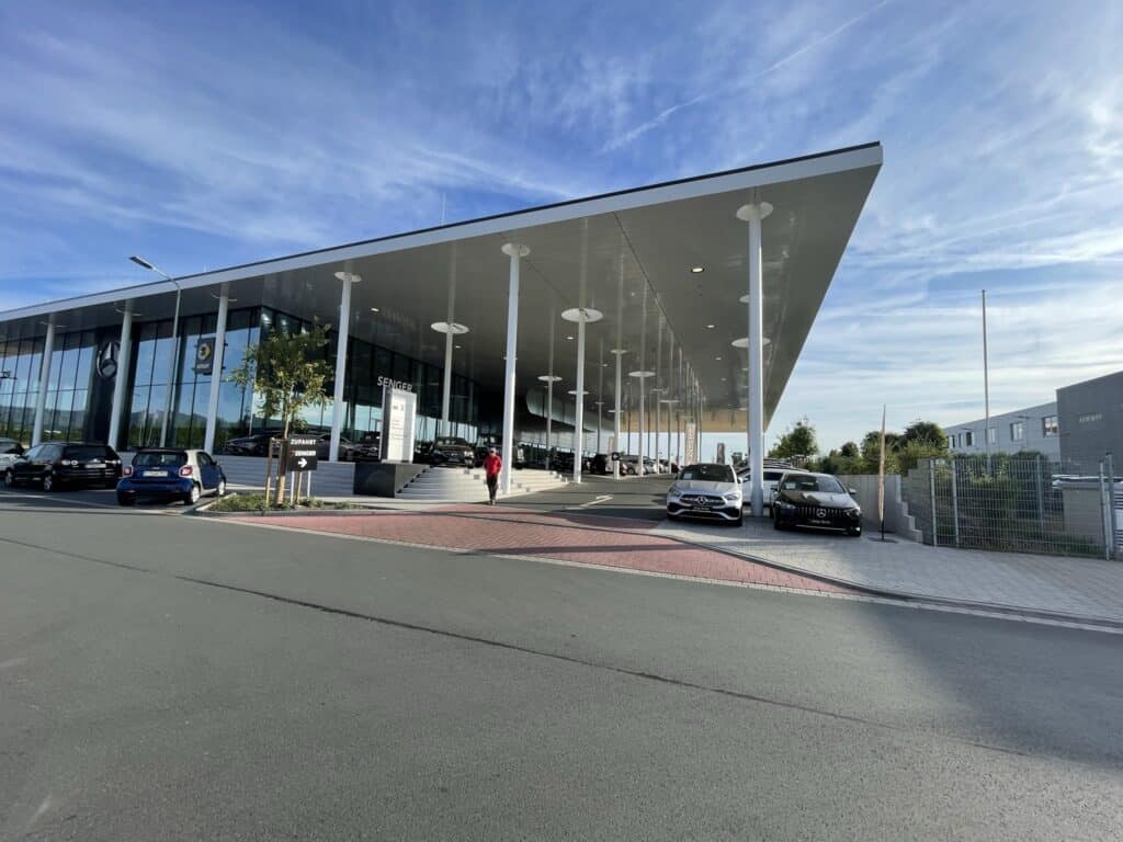 Seitliche Perspektive des Autohauses Senger in Oberursel mit weit auskragendem Dach, schlanken Stützen und geparkten Fahrzeugen.
