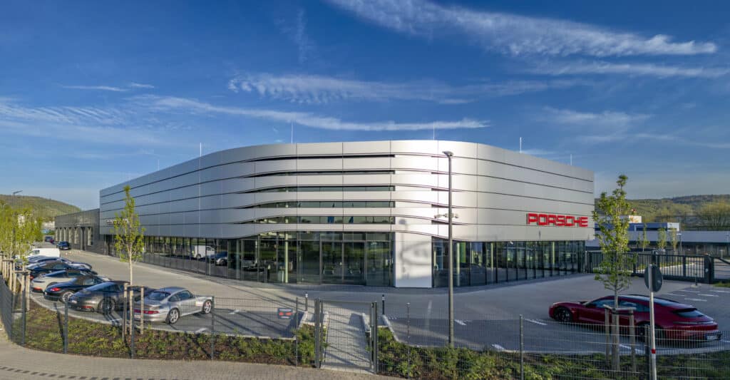 Außenansicht des Porsche Zentrums Karlsruhe mit moderner Aluminiumfassade und geparkten Porsche-Fahrzeugen bei blauem Himmel.