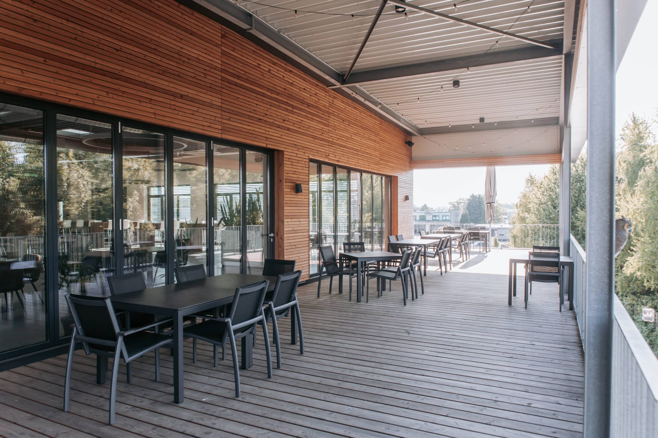 Überdachte Außenterrasse der Kantine mit Holzboden, Tischen und Stühlen und Blick ins Grün.
