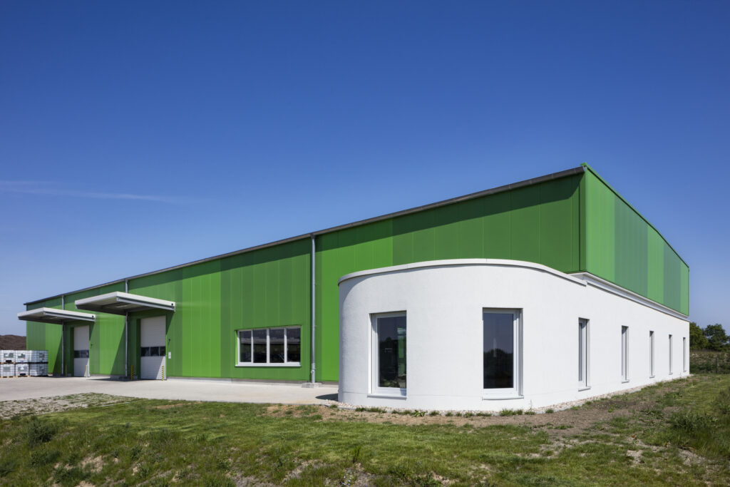 Außenansicht einer neu errichteten Lager- und Produktionshalle in Mettmann mit markanter grüner Fassadenverkleidung und vorgelagertem weißem Bürotrakt unter blauem Himmel.