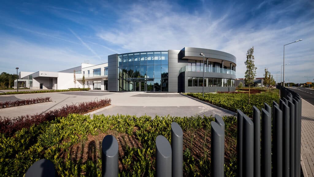 Modernes Firmengebäude mit großer, gebogener Glasfassade im Eingangsbereich und angrenzenden weißen Gebäudeteilen. Im Vordergrund befinden sich gepflegte Grünflächen und eine Reihe dunkler, vertikaler Zaunelemente entlang des Gehwegs. Der Himmel ist klar und blau.