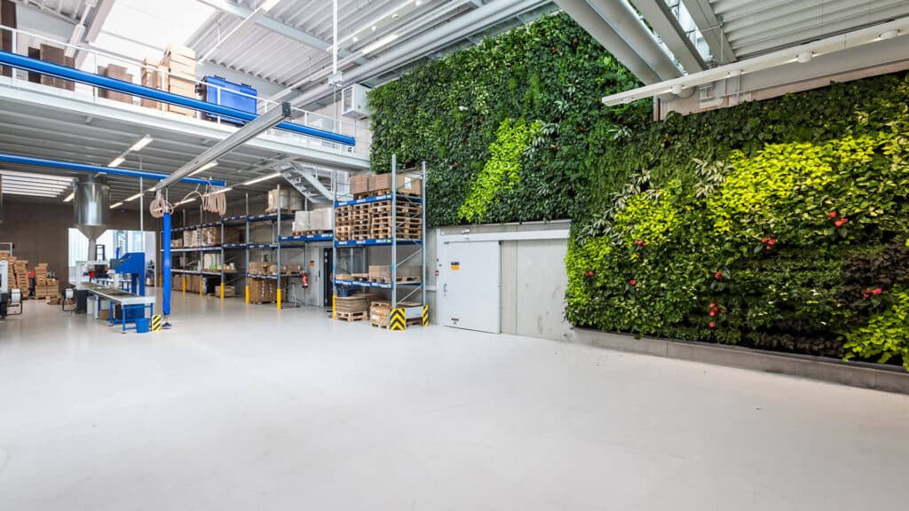 Moderne Lager- und Produktionshalle mit grüner Pflanzenwand, Hochregalen, Arbeitsstationen und offener, heller Industriearchitektur.
