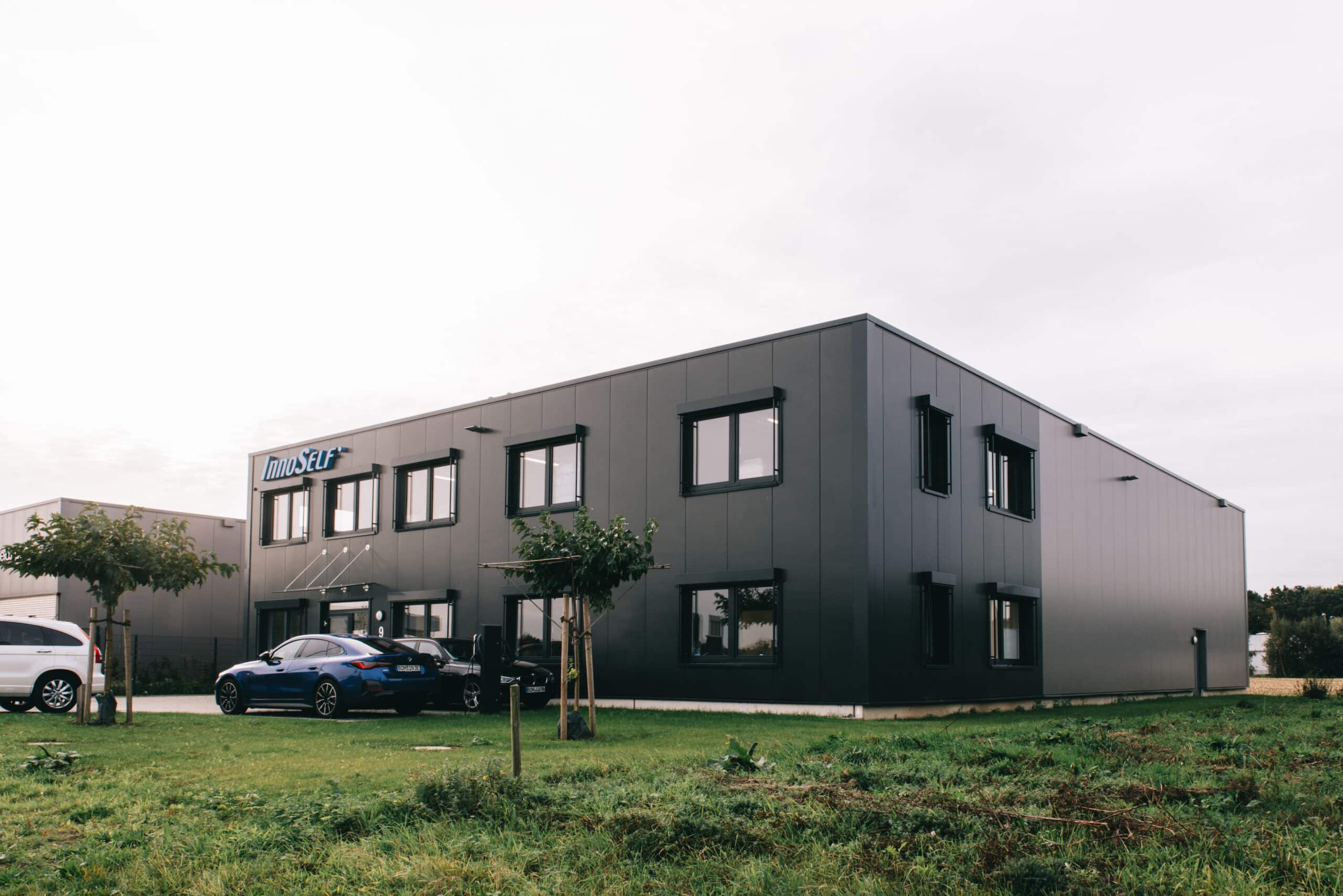 Seitenansicht des InnoSelf-Gebäudes mit Parkplatz, Grünfläche und moderner Fassadengestaltung.