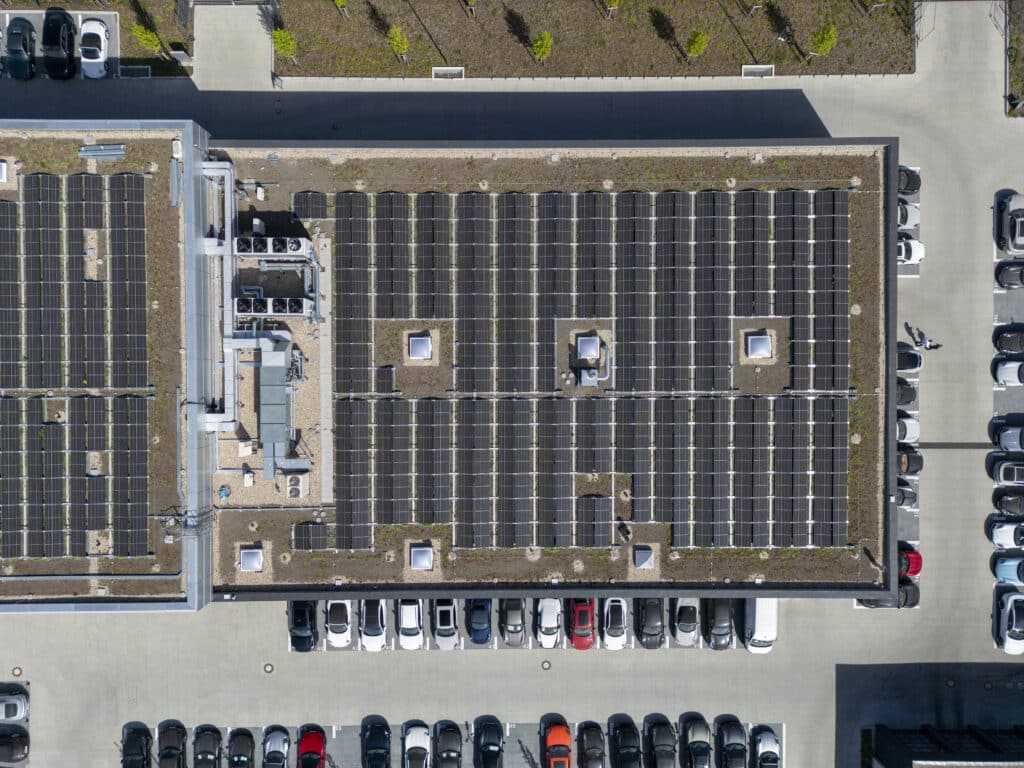 Drohnenaufnahme des Porschezentrums, die das große Flachdach mit einer vollflächig installierten Photovoltaikanlage zeigt. Die Solarmodule sind in gleichmäßigen Reihen angeordnet; in der Mitte befinden sich mehrere technische Aufbauten und Dachöffnungen. Rund um das Gebäude stehen zahlreiche geparkte Autos.