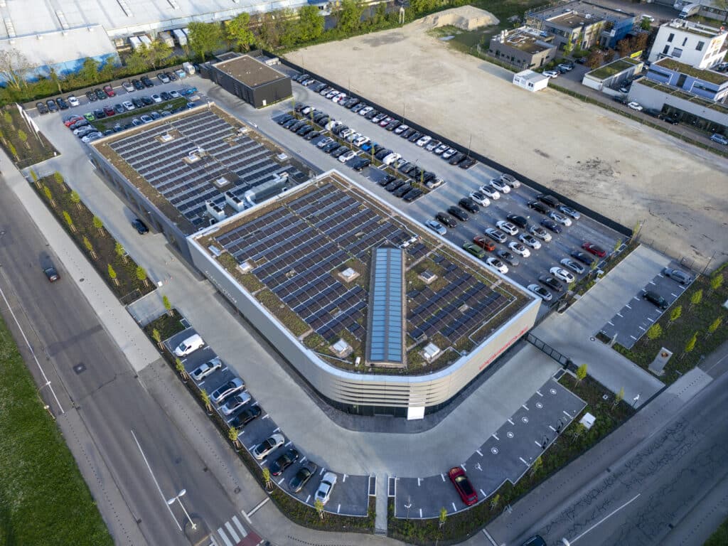 Drohnenaufnahme des Porschezentrums mit zwei großflächigen Photovoltaikanlagen auf den begrünten Flachdächern. Das Gebäude ist von zahlreichen Parkplätzen umgeben, viele davon belegt. Die Anlage liegt an einer Hauptstraße, angrenzend an Gewerbeflächen und freie Grundstücke.