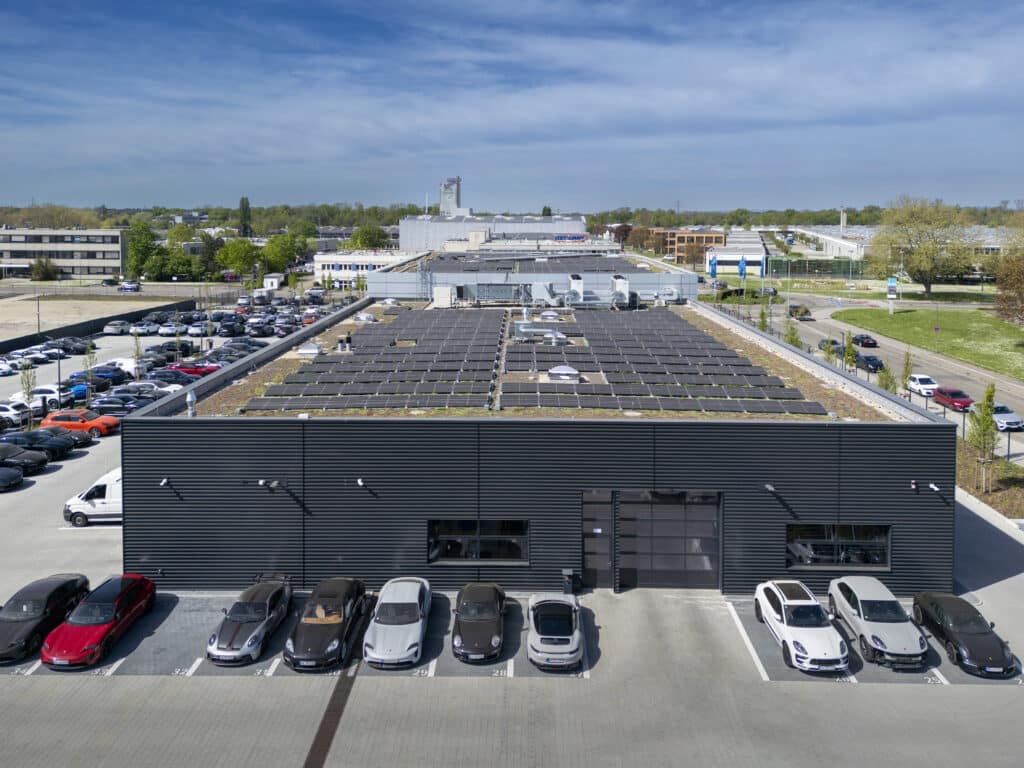 Drohnenaufnahme des Porschezentrums, die das große Flachdach mit einer vollflächig installierten Photovoltaikanlage zeigt. Die Solarmodule sind in gleichmäßigen Reihen angeordnet; in der Mitte befinden sich mehrere technische Aufbauten und Dachöffnungen. Rund um das Gebäude stehen zahlreiche geparkte Autos.
