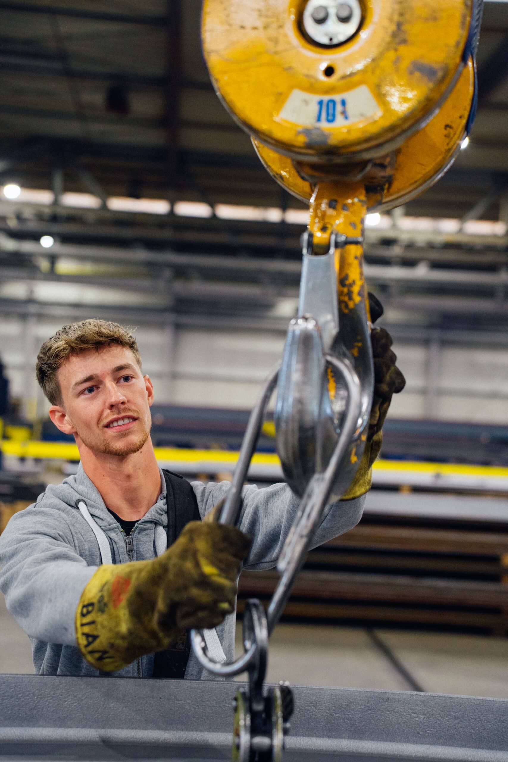 Ein Arbeiter in einer Werkhalle befestigt mit Handschuhen einen Metallhaken an einem gelben Lastkran.