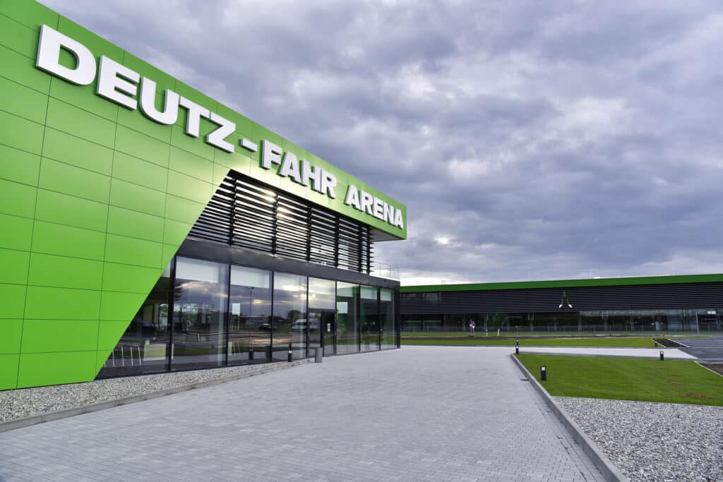 Nahaufnahme der modernen Deutz-Fahr Arena mit ihrer markanten grünen Fassade und großen Glasfronten. Der Vorplatz ist gepflastert, rechts schließt eine gepflegte Rasenfläche an. Über dem Haupteingang prangt deutlich der Schriftzug ‚DEUTZ-FAHR ARENA‘.