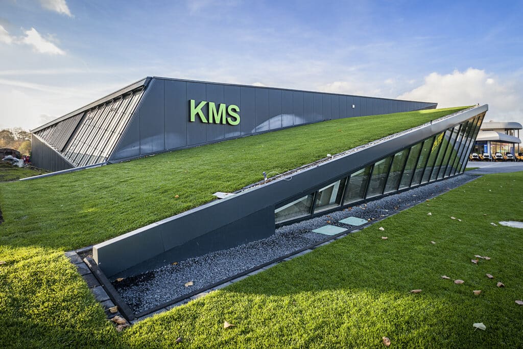 Moderne Ausstellungshalle in Kamp-Lintfort mit markant geneigtem, begrüntem Dach und großzügigen Glasflächen. Die Fassade besteht aus grauen Metallpaneelen, auf denen der grüne Schriftzug ‚KMS‘ hervorsticht. Die Architektur zeichnet sich durch ihre dynamische, schräge Form und die teilweise eingegrabene Glasfront aus, die viel Tageslicht ins Gebäude lässt.