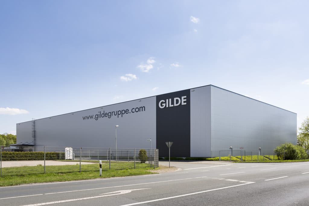 Außenansicht eines neu errichteten Hochregallagers in Bocholt mit weiß-grauer Sandwichpaneel-Fassade, klaren vertikalen Linien und großem Firmenlogo ‚GILDE‘. Die Halle verfügt über eine extrem hohe Bauweise für automatische Hochregalsysteme. Im Vordergrund liegen eine umlaufende Zaunanlage, eine Zufahrtsstraße und begrünte Außenflächen bei sonnigem Wetter.