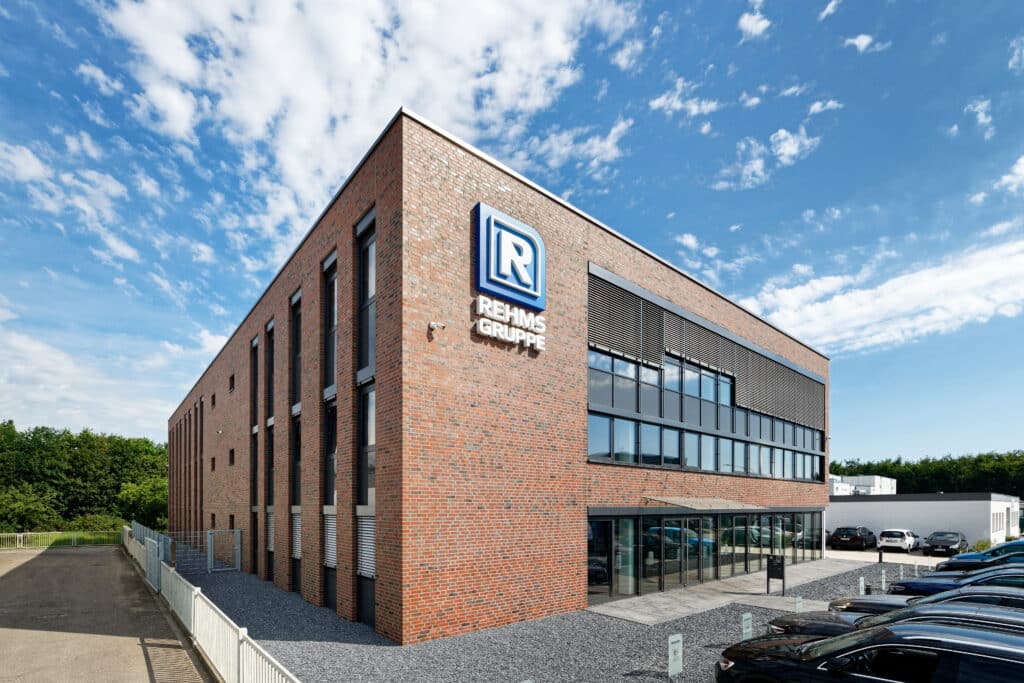 Backsteinernes Schulungszentrum der Rehms-Gruppe mit moderner Glasfassade und markantem Firmenlogo. Vor dem Gebäude befinden sich Parkplätze, der Himmel ist blau mit leichten Wolkenmustern.