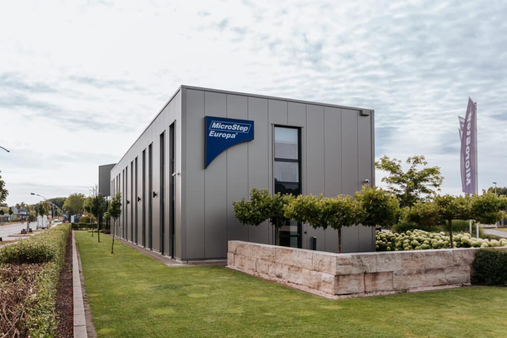 Gewerbegebäude von MicroStep Europa mit einer Fassade aus vertikal montierten Sandwich-Wandelementen in grauer Metalloptik. Die schmalen, hohen Fensterstreifen gliedern die Fassade rhythmisch. Davor liegen gepflegte Grünflächen mit niedrigen Formbäumen, Hecken und einer Natursteinmauer, während im Hintergrund Firmenflaggen und eine Straße zu sehen sind.