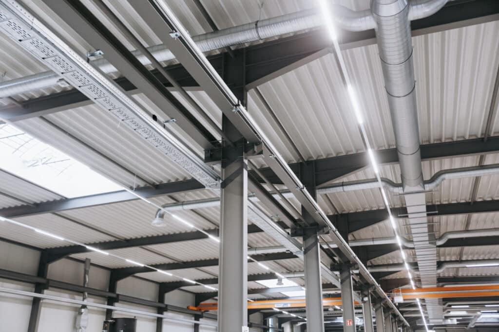 Industriehalle mit Stahlträgern, sichtbaren Lüftungsleitungen und moderner LED-Deckenbeleuchtung.