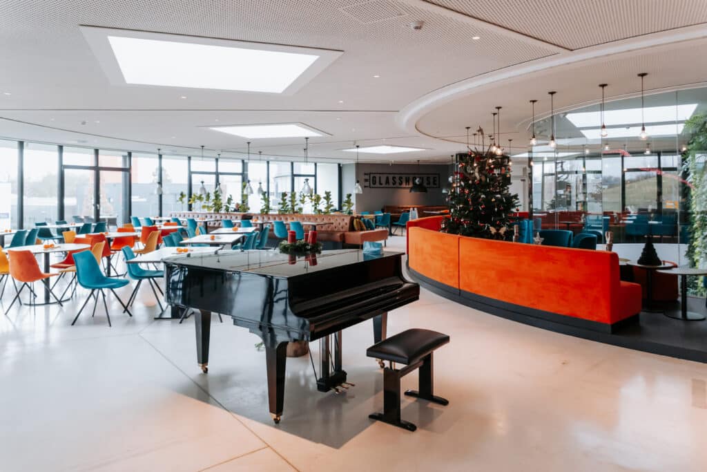 Moderner Lounge- und Gastroraum mit schwarzem Flügel, orangefarbener Rundsitzbank, Weihnachtsbaum und farbigen Stühlen.