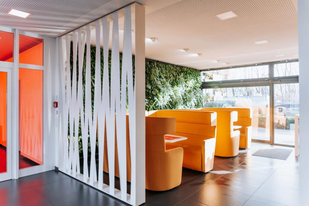 Moderner Loungebereich mit orangefarbenen Sitznischen, grüner Pflanzenwand und großen Glasfenstern.