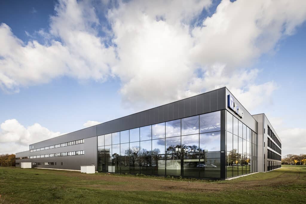 Moderne, langgestreckte Büro- und Forschungsgebäude-Fassade mit großflächigen Glasfronten und dunklen Metallpaneelen eines R&D-Centers. Die spiegelnden Fenster reflektieren Bäume und den Himmel, während sich das Gebäude klar von der umliegenden Grünfläche abhebt. Unter einem lebhaften Wolkenhimmel präsentiert sich die Architektur geradlinig, technisch und repräsentativ.