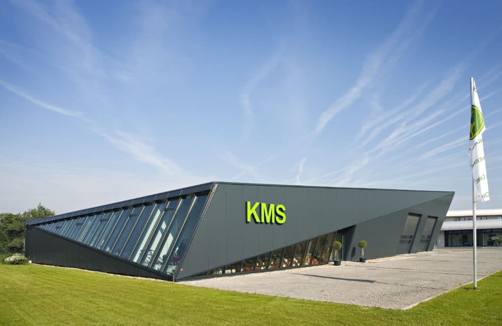 Moderne Ausstellungshalle mit markanter, schräg verlaufender Glasfassade und dunkelgrauen Metallpaneelen. Auf der Frontseite befindet sich ein großes grünes „KMS“-Logo. Der Baukörper ist dynamisch, keilförmig und liegt auf einer gepflegten Rasenfläche, daneben ein Vorplatz mit Kies. Rechts steht eine Flagge im Wind vor blauem Himmel mit leichten Wolkenstreifen.