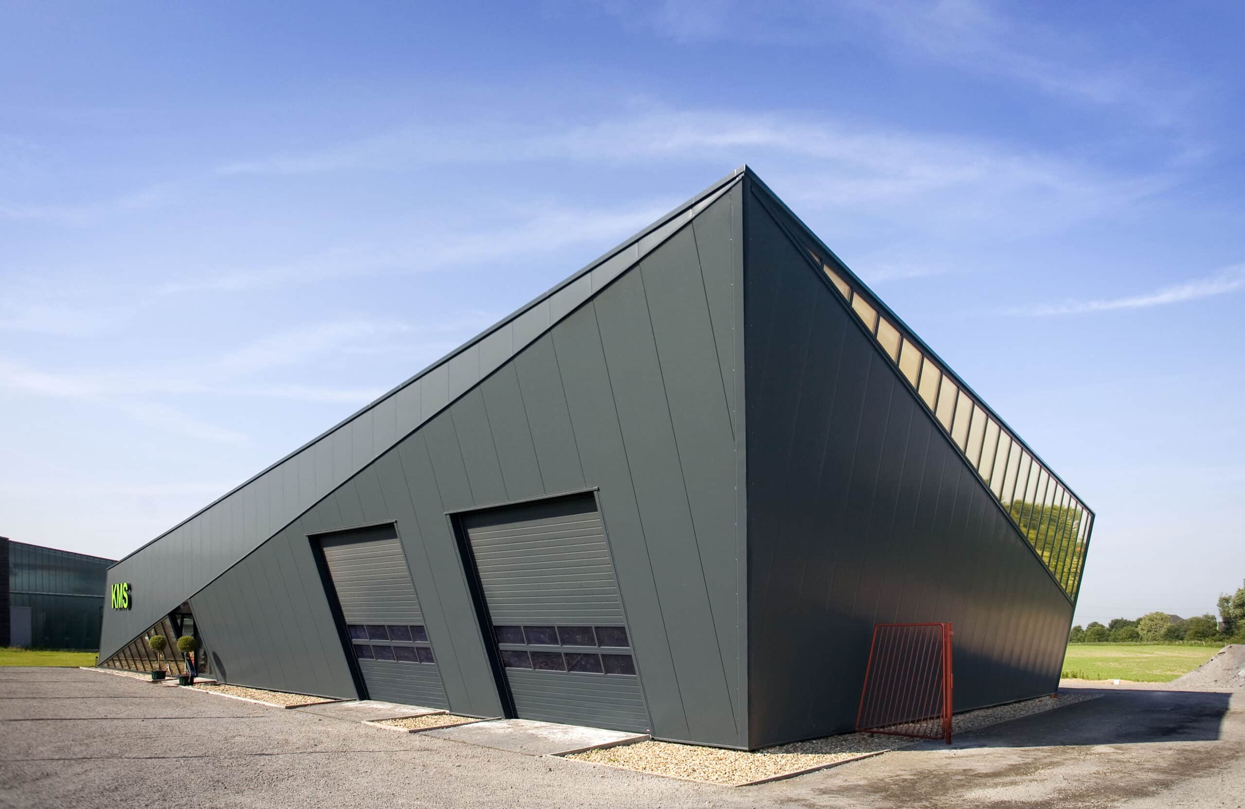 Schräg und keilförmig gestaltete Ausstellungshalle mit dunkelgrauen Metallpaneelen. Zwei geneigte Sektionaltore befinden sich an der Längsseite. Entlang der rechten Dachkante verläuft ein langes, schmales Fensterband. Die moderne, dynamische Architektur steht frei auf einer Kiesumrandung, dahinter weitläufige Grünflächen unter klarem, blauem Himmel.